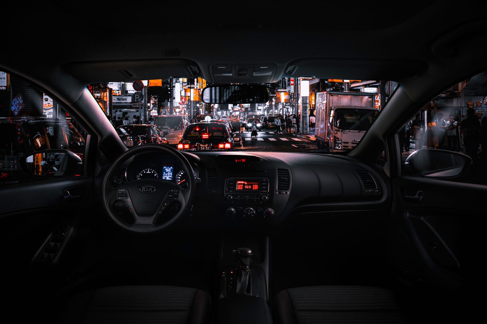 Night driving scene showing a dark car interior and steering wheel silhouette with a brightly lit city street ahead