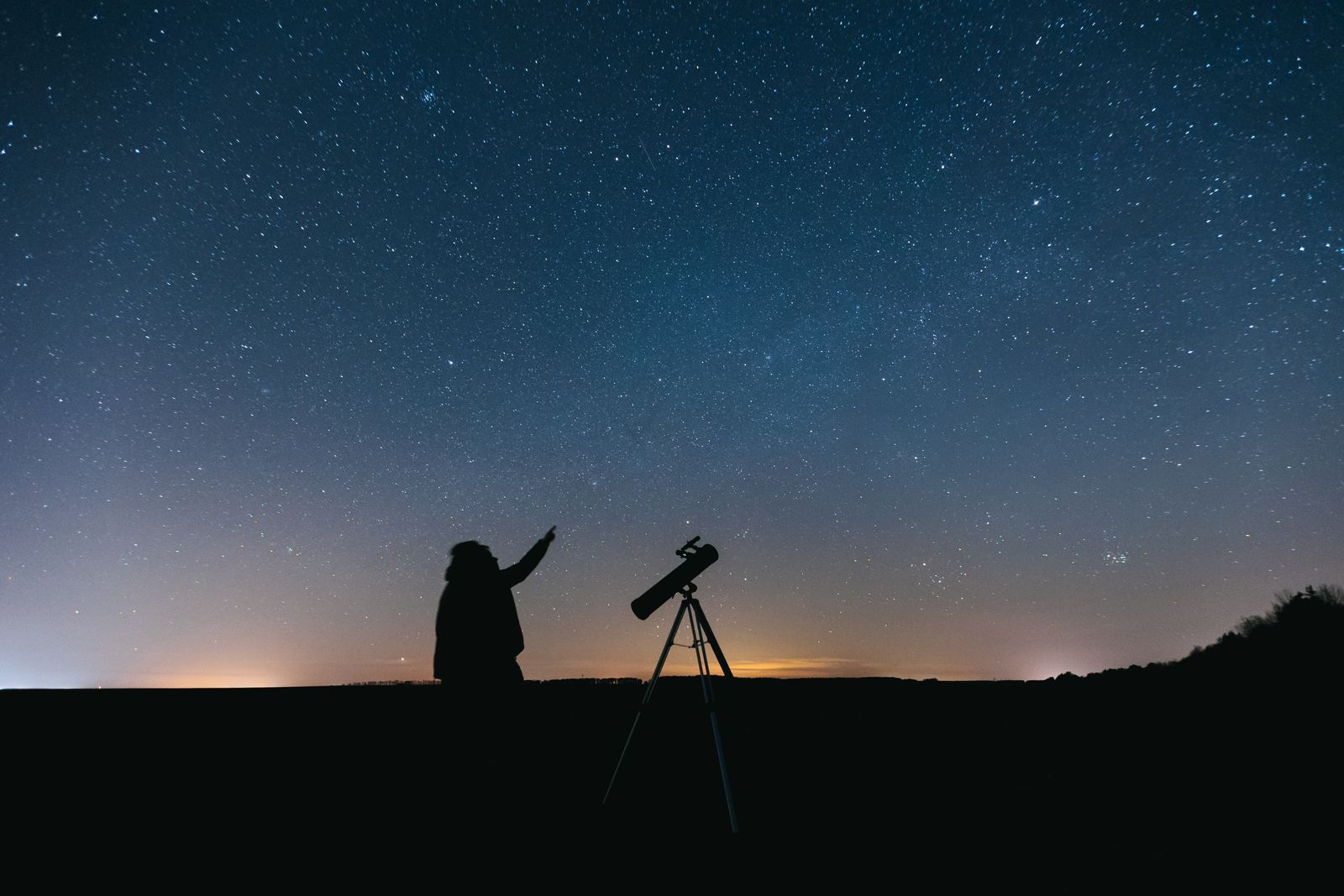 Silhouette of a person standing beside a telescope under a star-filled sky
