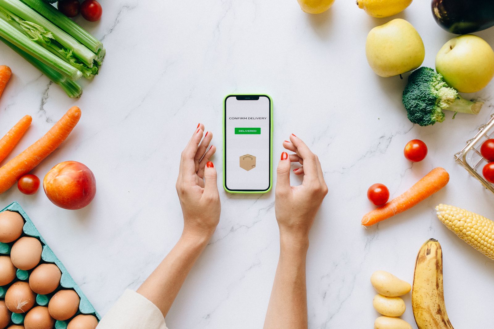 Person holding a smartphone displaying a food or grocery app near a spread of fresh vegetables and fruit