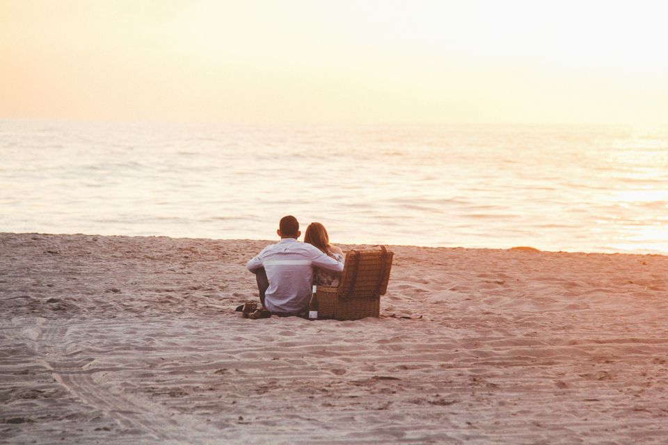 Couple sitting on a sandy beach at sunset with a cooler nearby for their picnic