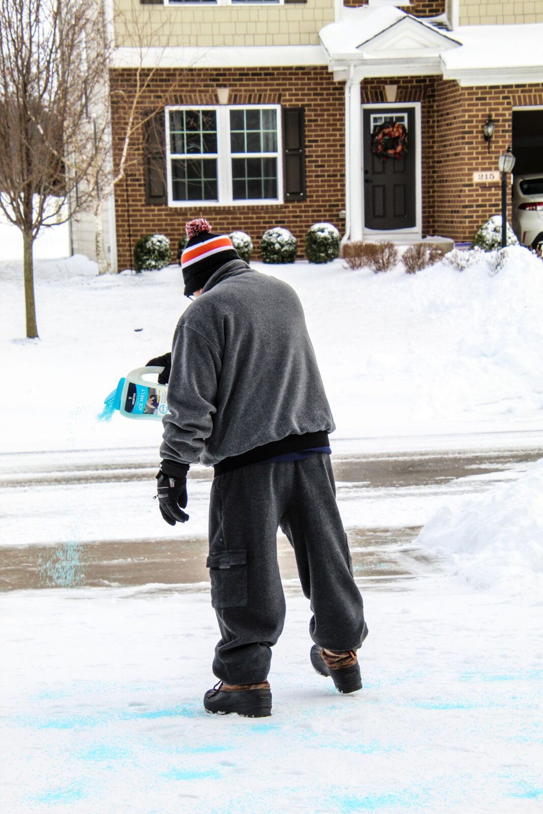 Person applying ice melt granules to a snow-covered driveway outside a home