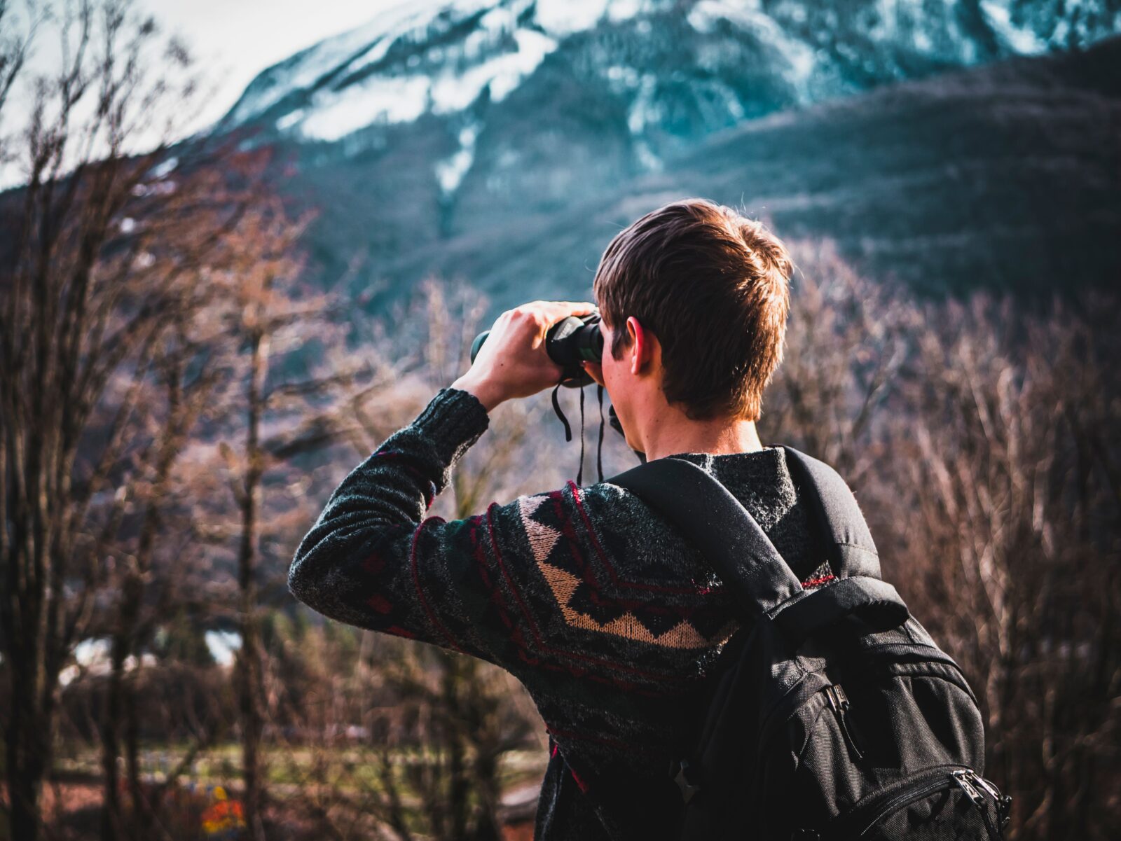 A man wearing a backpack holds binoculars to his eyes, looking out over a mountainous, wooded landscape.
