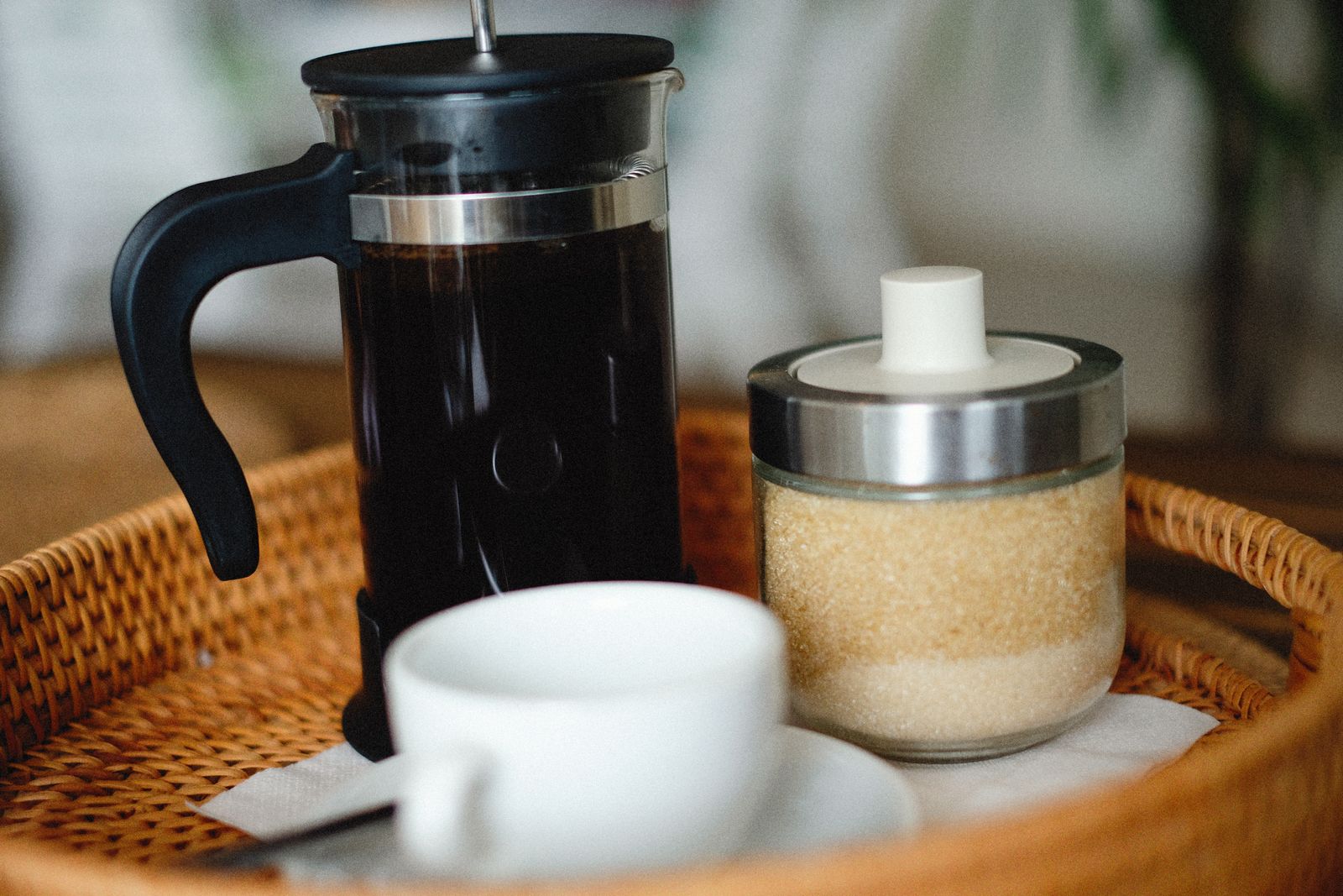 A French press, sugar jar, and coffee cup arranged on a wicker tray