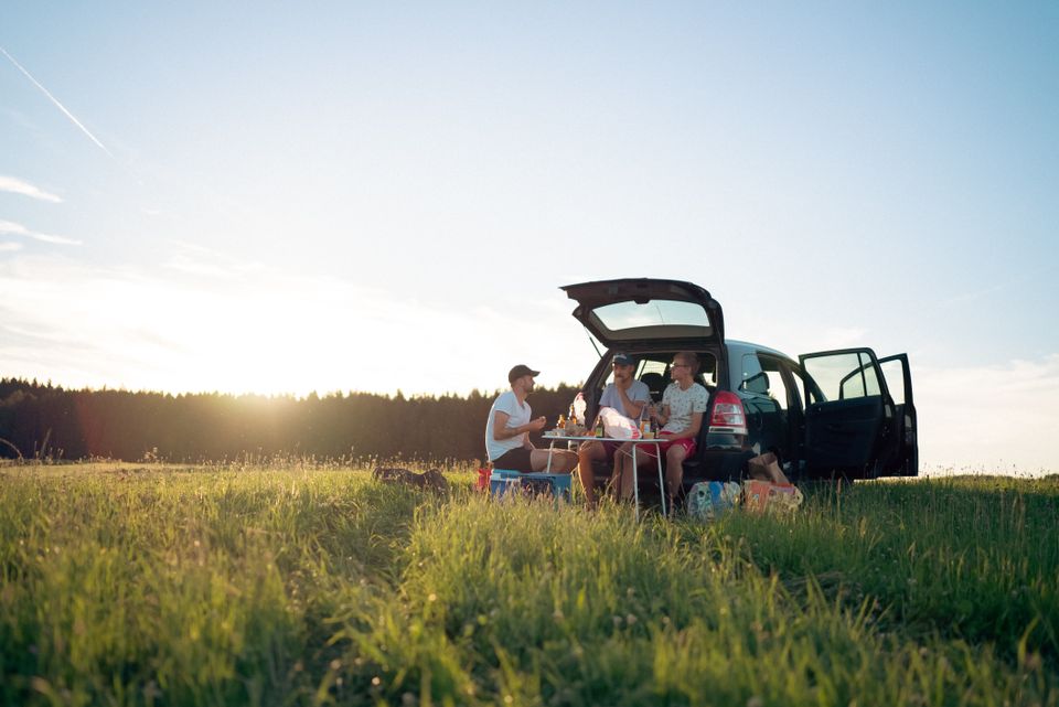 Friends gathered around a car's open trunk for a picnic with drinks and snacks at sunset