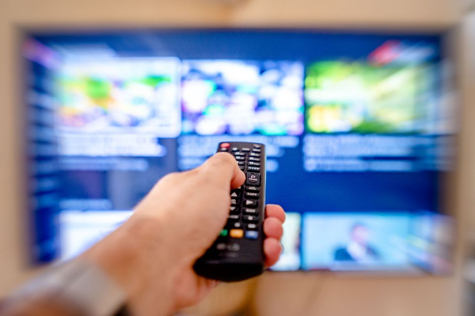 Close-up of a hand holding a black TV remote control pointed at a blurred television screen