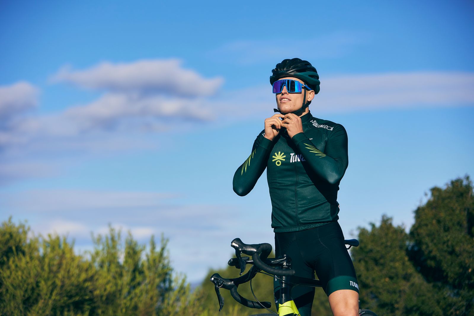 Male cyclist standing outdoors in sportswear adjusting his helmet before a ride, rural landscape in the background