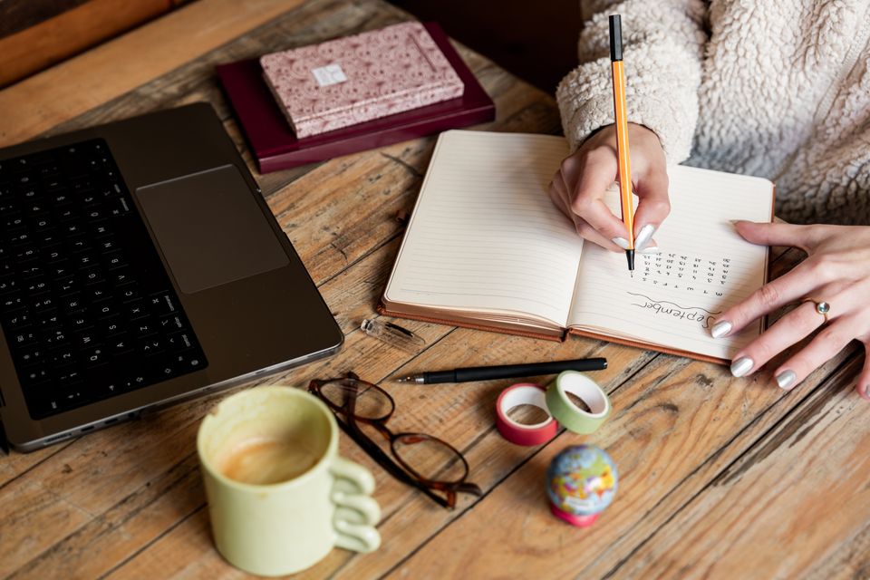 Woman writing in a notebook at a wooden desk with a laptop and coffee cup nearby