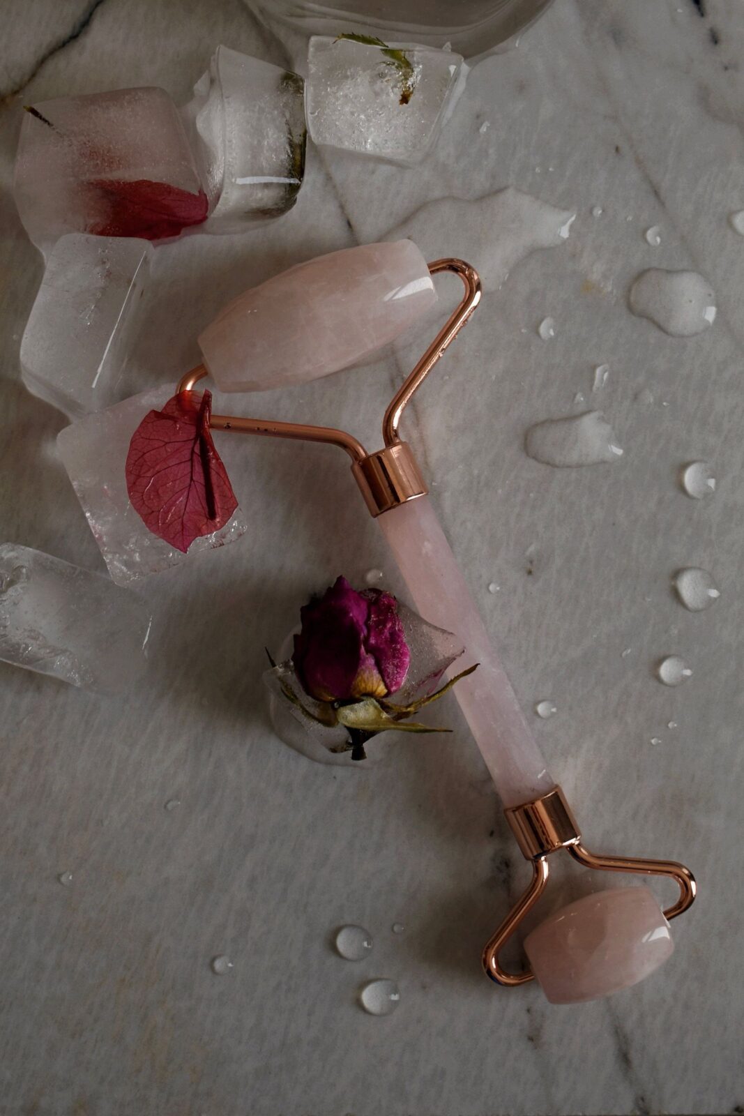 Rose quartz facial roller resting beside ice cubes and rose petals on a marble surface