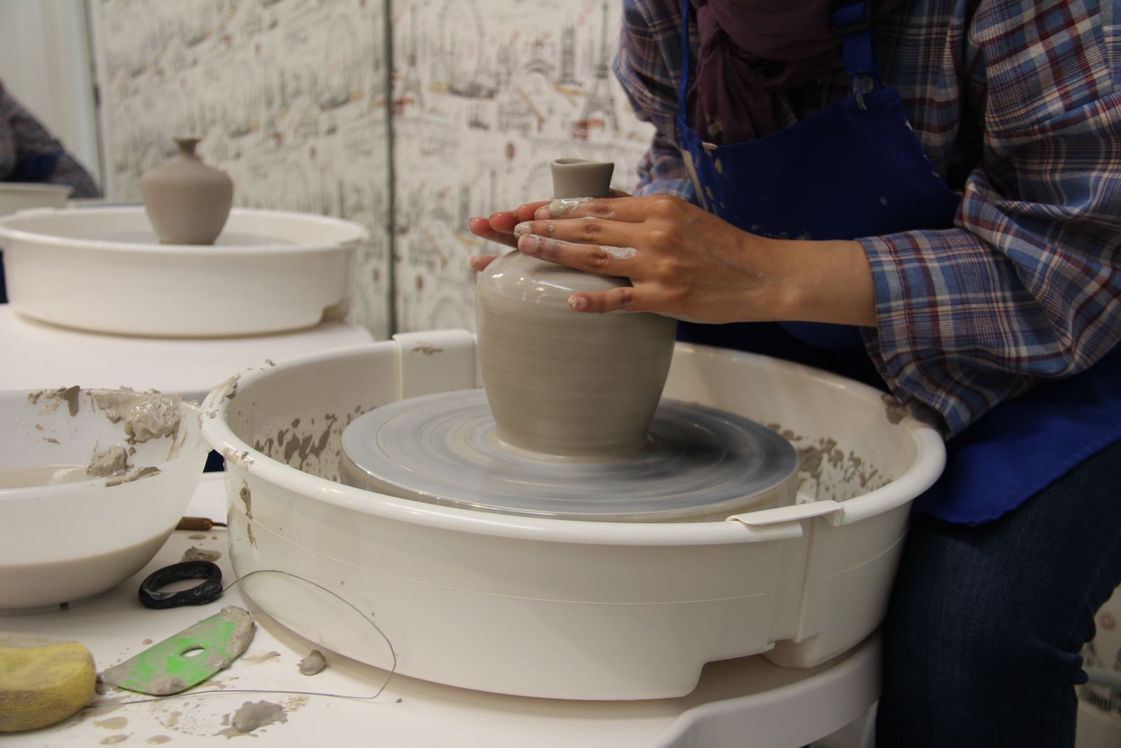 Hands shaping wet clay into a tall vase form on a pottery wheel in a small studio