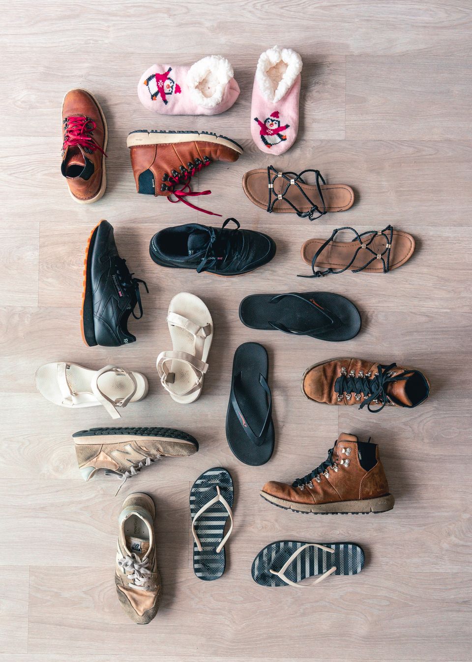 A mixed pile of sneakers, boots, sandals, and slippers arranged on a wooden floor