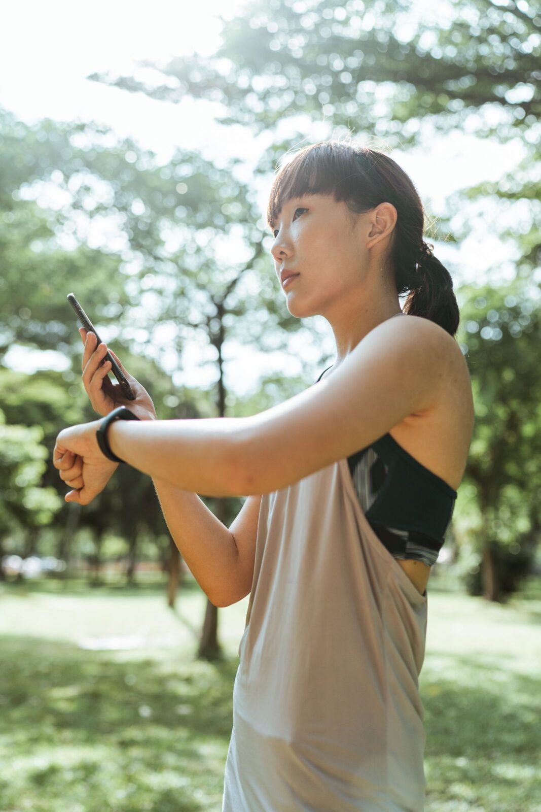 A woman outdoors glancing at a wrist wearable while holding her phone, representing checking synced health data in a companion app