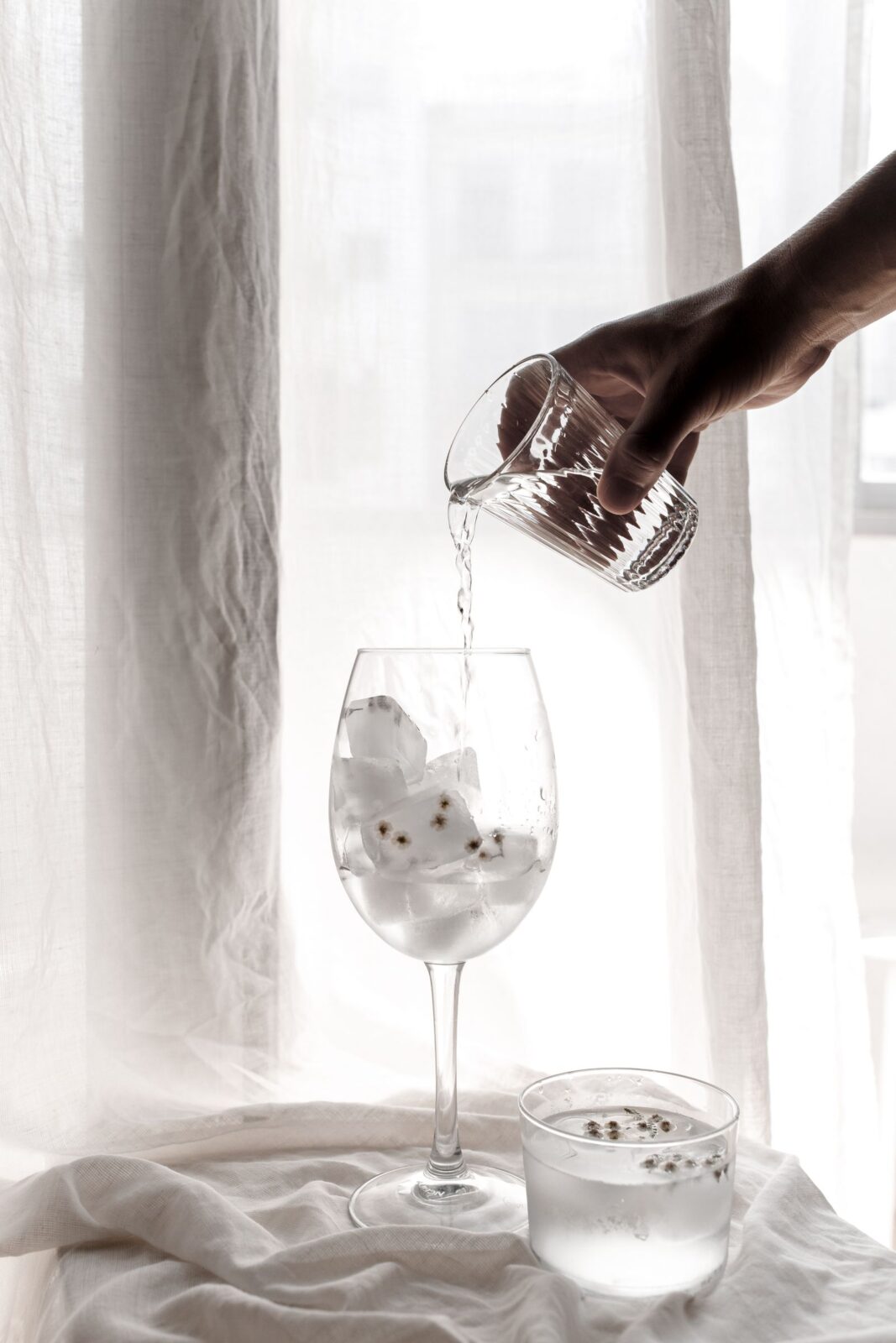 A hand pouring a clear liquid over large ice cubes in a stemmed glass