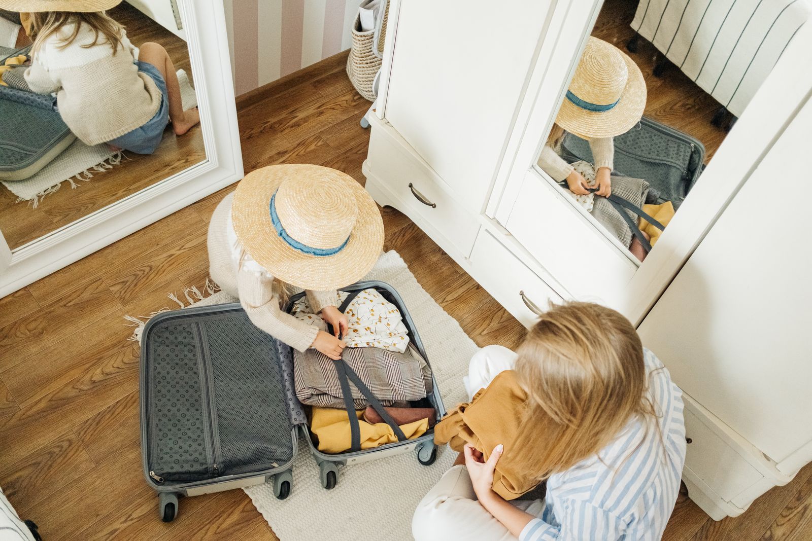 A mother and young child packing folded clothes into an open suitcase on the floor before a family trip