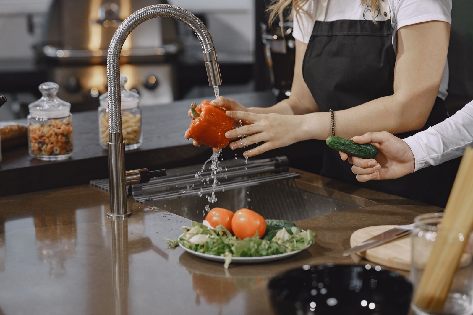 A person washing a red bell pepper and other vegetables at a kitchen sink