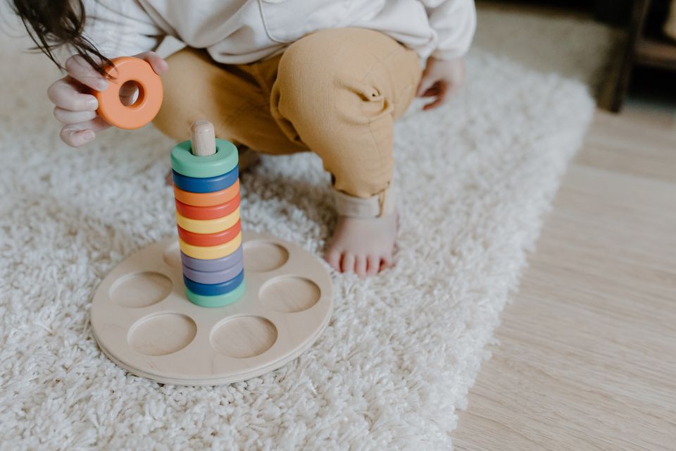 Toddler's hands stacking colorful wooden rings on a wooden ring stacker toy while seated on a carpet