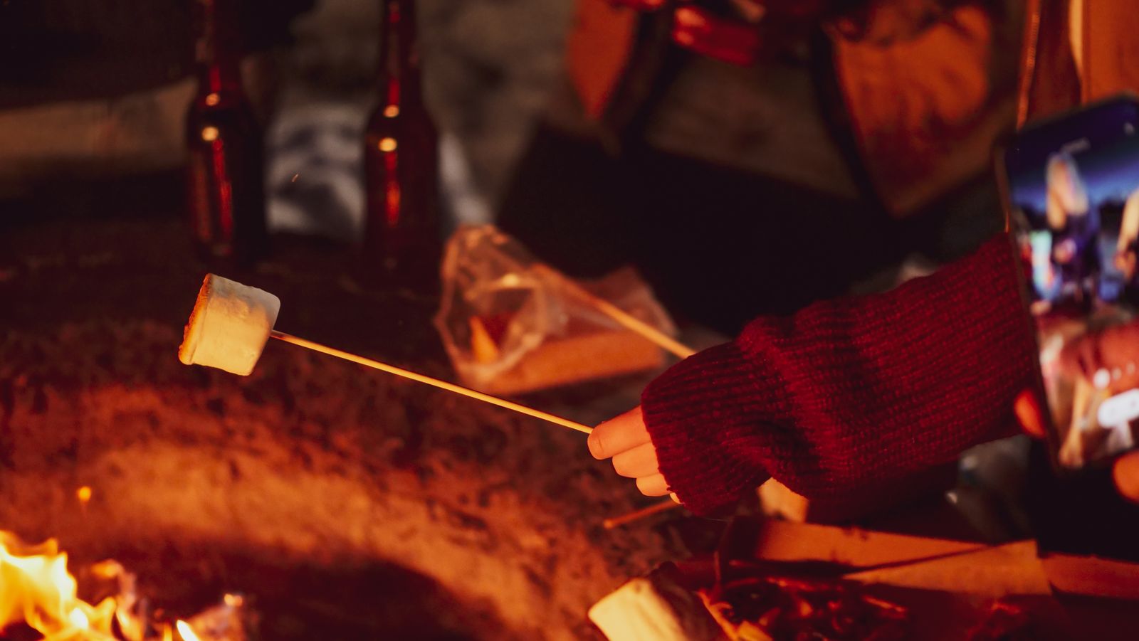A group of friends gathered around an outdoor fire pit roasting marshmallows on a fall evening