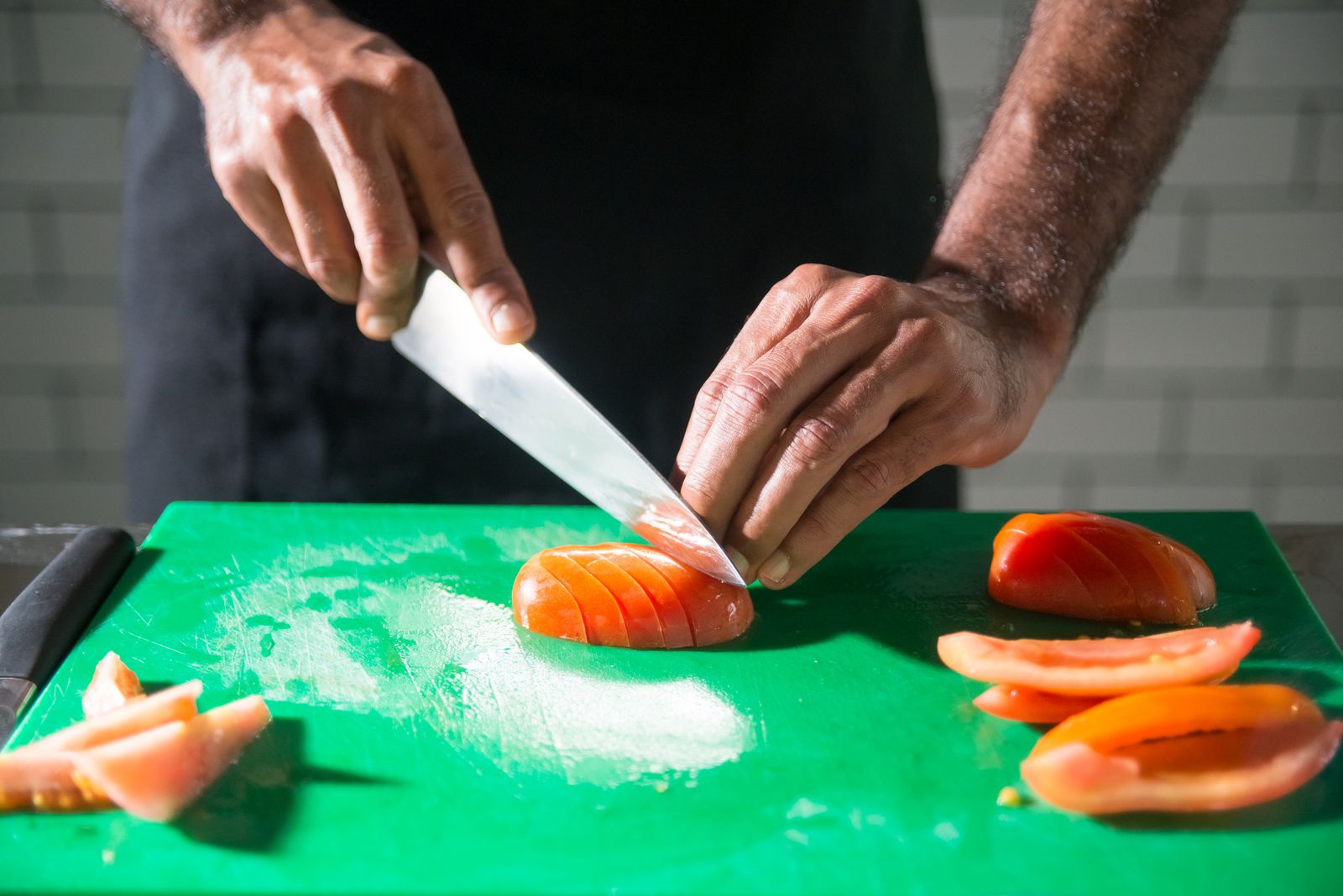 A hand carefully guiding a tomato across a cutting board while slicing