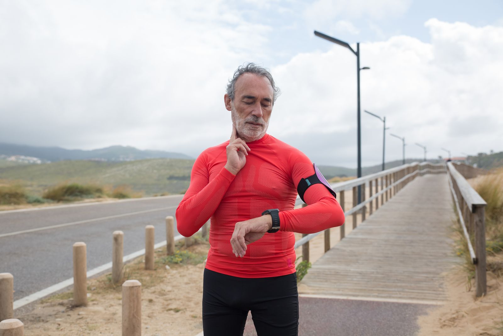 Older man in activewear checking his heart rate on a smartwatch along a coastal jogging path