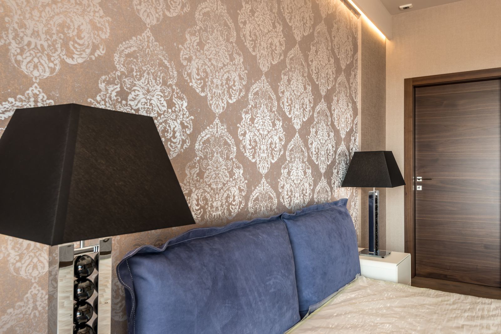 A bedroom wall covered in a silver damask-pattern wallpaper behind a blue headboard and black lamps