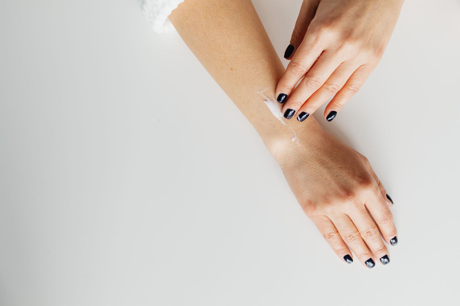 Two hands spreading white cream along a forearm on a white surface