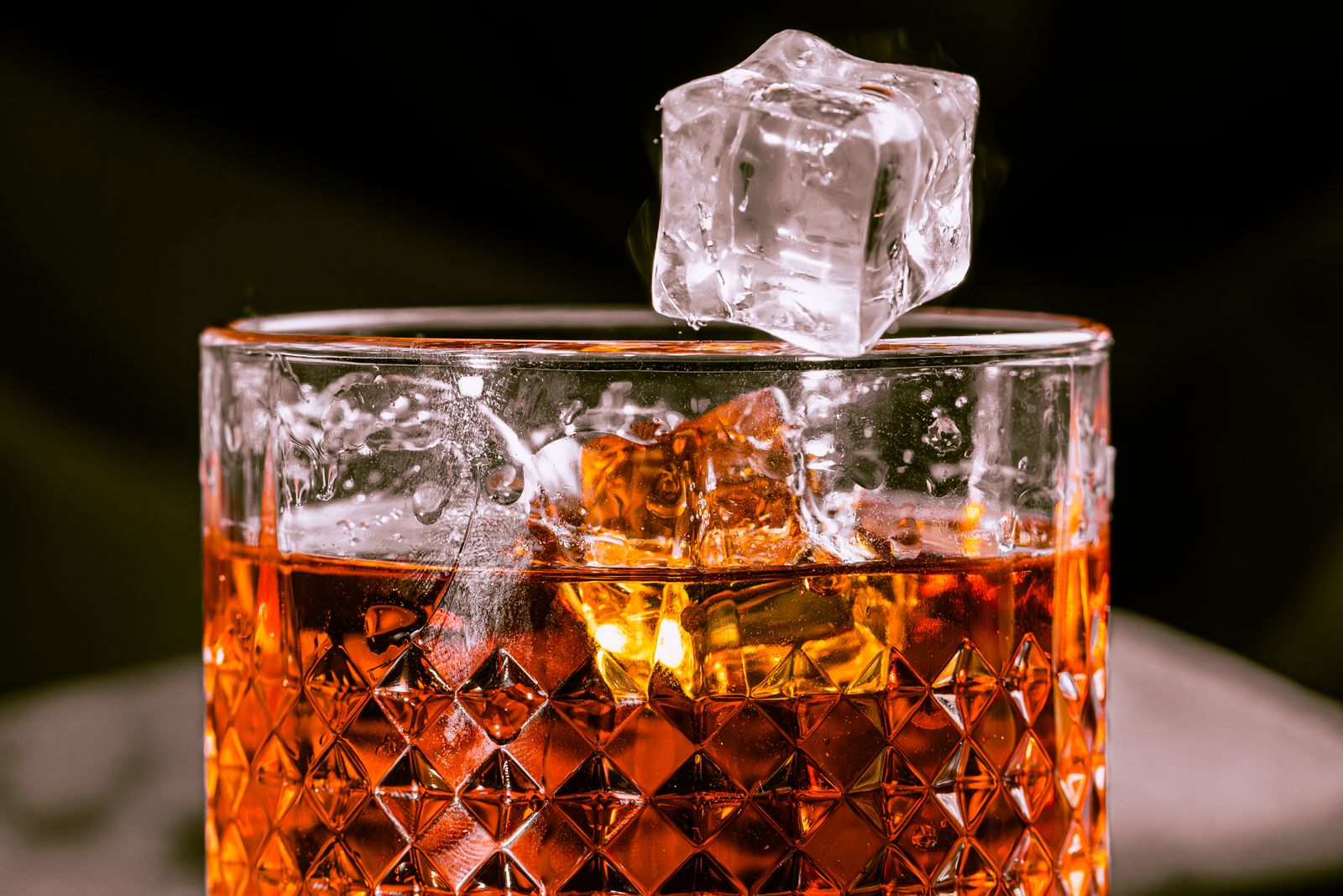 A clear ice cube resting on the rim of a cut-glass tumbler of whisky with more ice inside