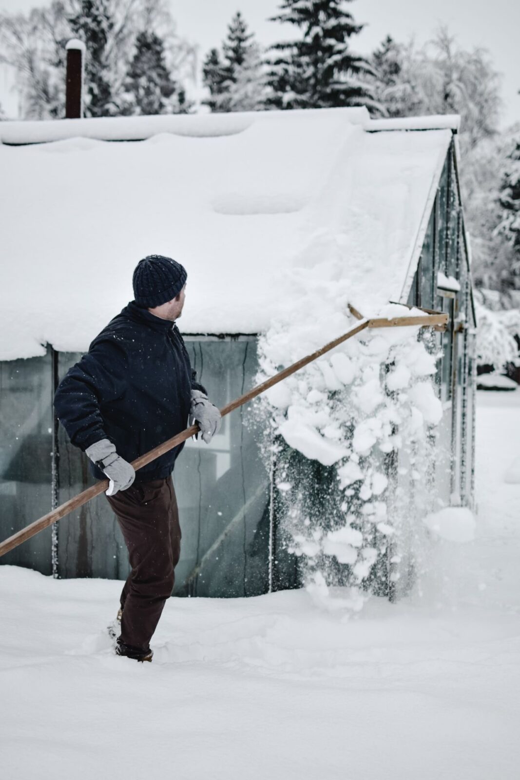 A man standing in deep snow pulling snow off a small building's roof with a long-handled rake