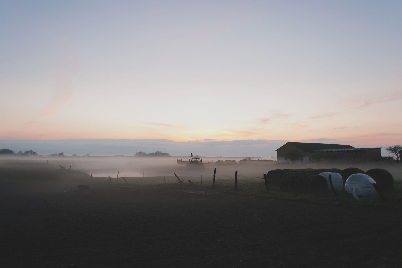 Misty farm field at dawn with a barn, round hay bales and fence posts under a pale orange sky