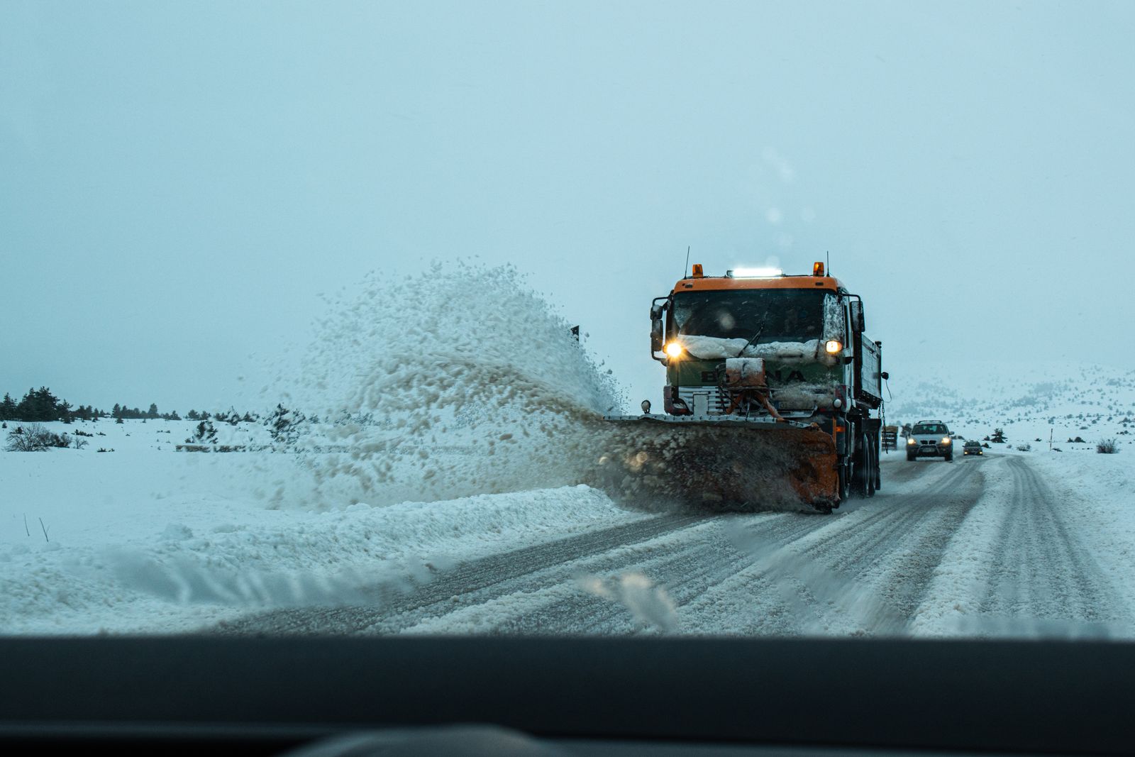A snowplow truck throwing a spray of snow off a rural road, seen through a car windshield