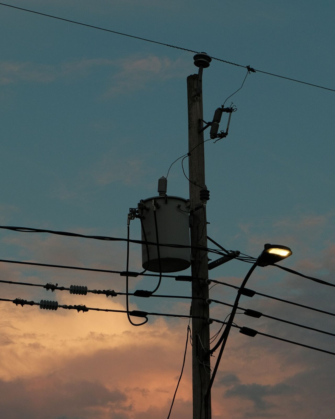 Utility pole with wires, a transformer and a street light silhouetted against a sunset sky
