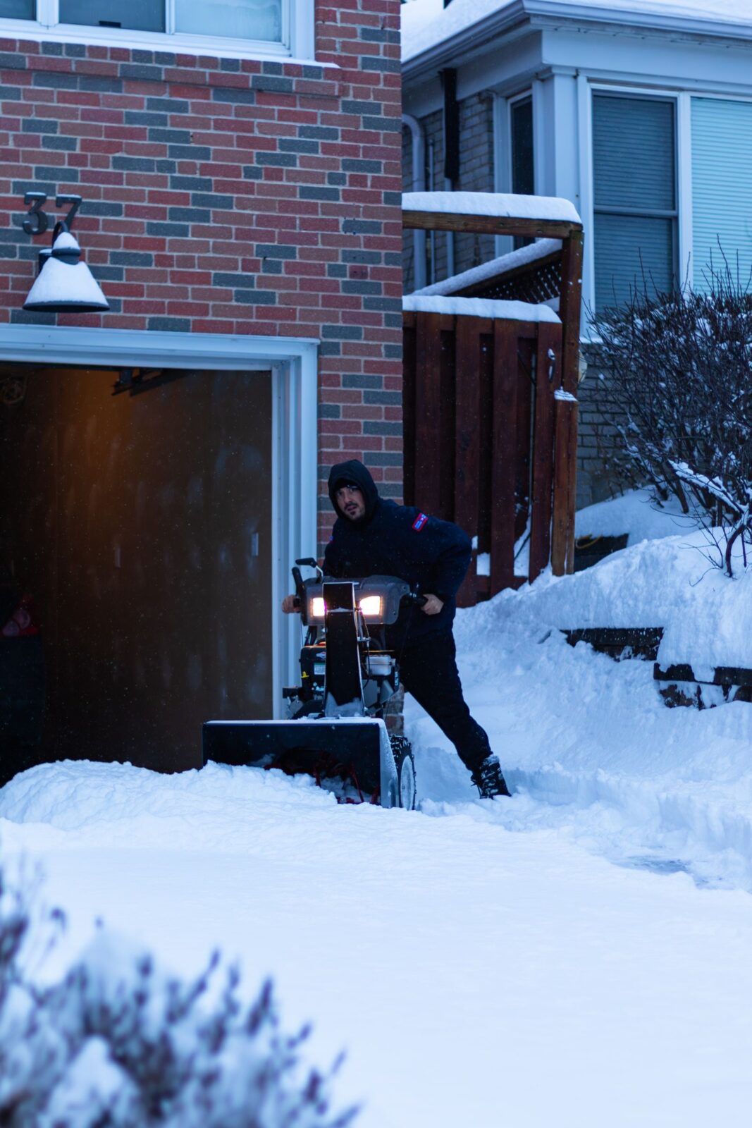 Snow blower on a driveway beside a brick house and open garage after a heavy snowfall