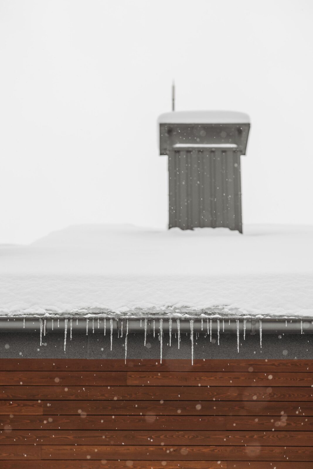 Snow-covered roof with a gray boxed chimney and icicles along the gutter during a winter snowfall
