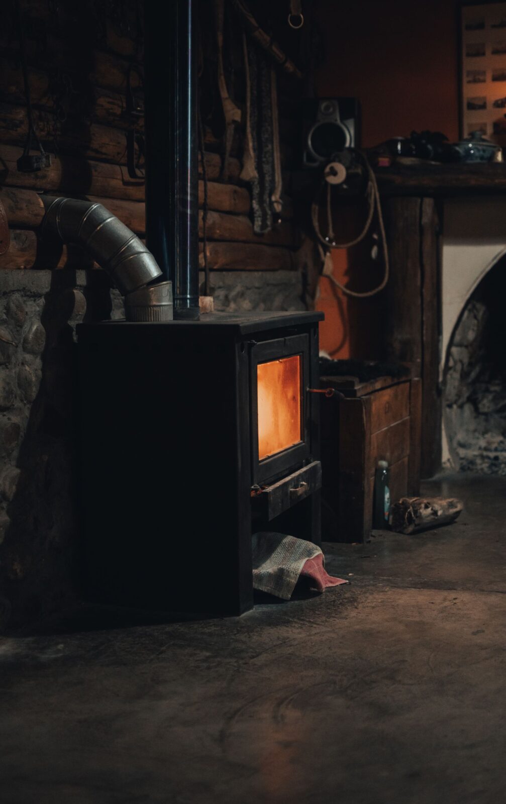 Black wood stove with a glowing fire and a stovepipe running to the wall of a log cabin