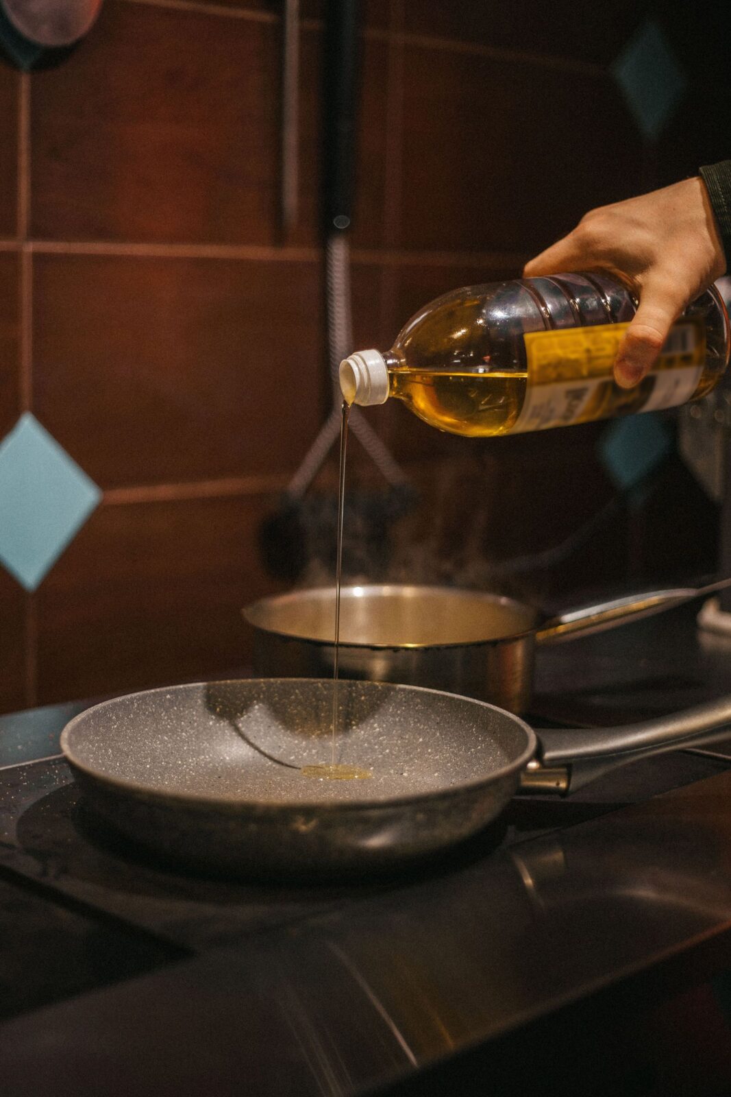 Hand pouring cooking oil from a bottle into a frying pan on a stove