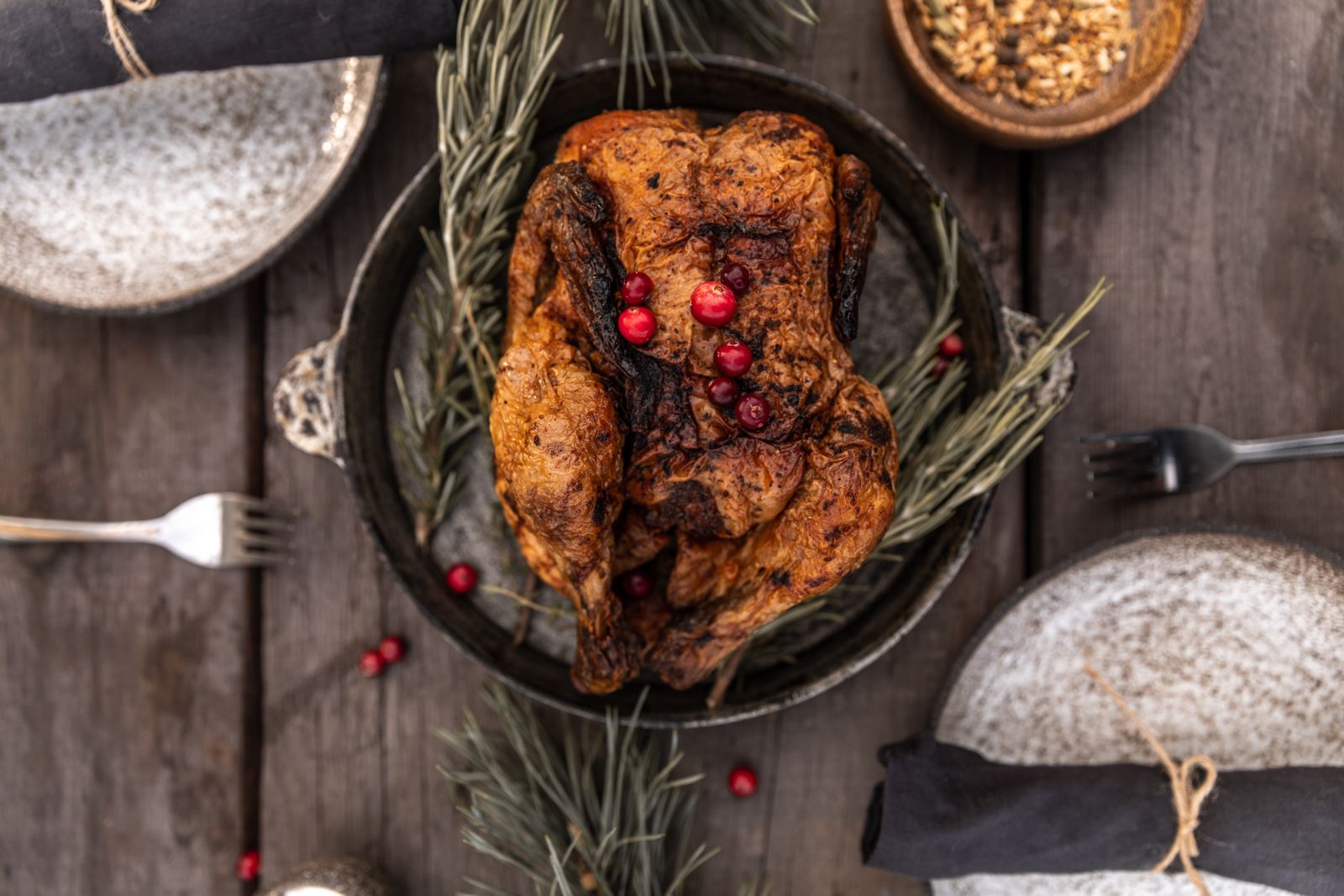 Whole roasted bird in a dark round pan with rosemary sprigs and cranberries on a wooden table