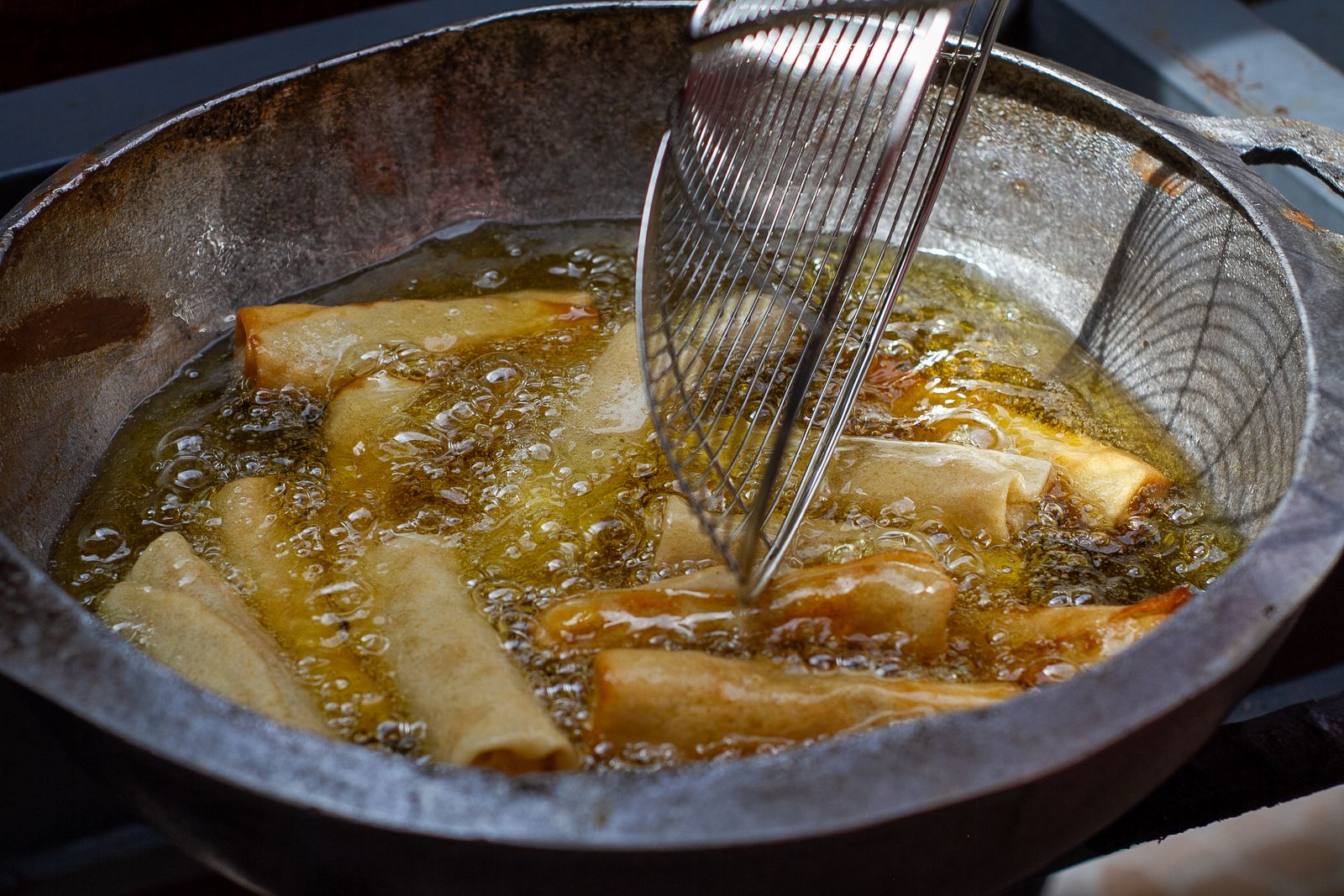 Spring rolls frying in hot oil with bubbles around them and a wire skimmer above the pot
